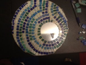 imageLarge round tilled mosaic with small circle mirror right centre. Small tiles follow the round shape in mixed colours green, blue and white. 